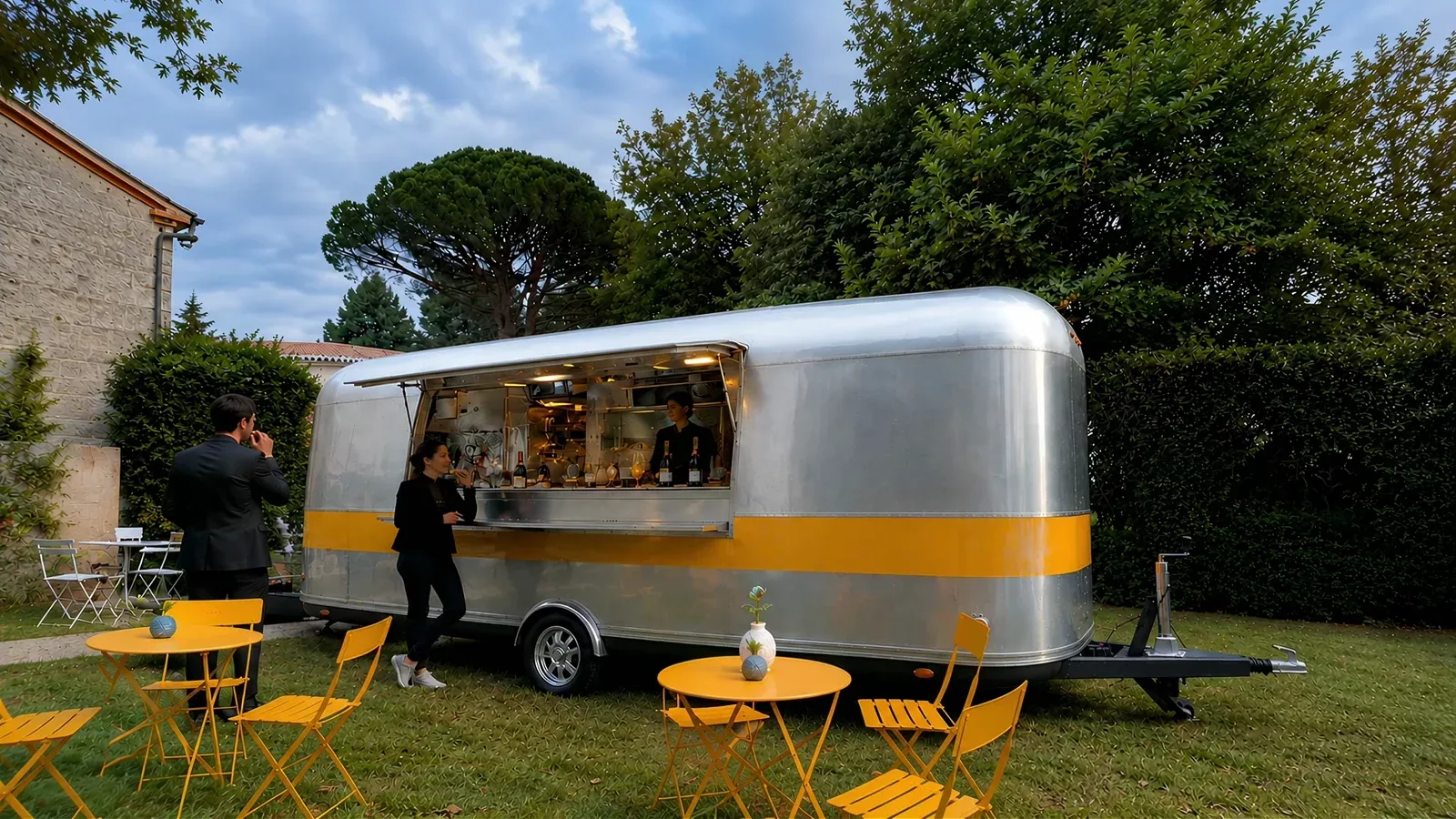 A silver food trailer serving customers in a garden